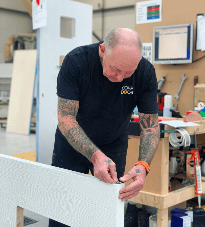 Bald, tattooed man in a "COMP door" t-shirt attaches a metal piece to a white wood-grain composite door frame in a workshop.