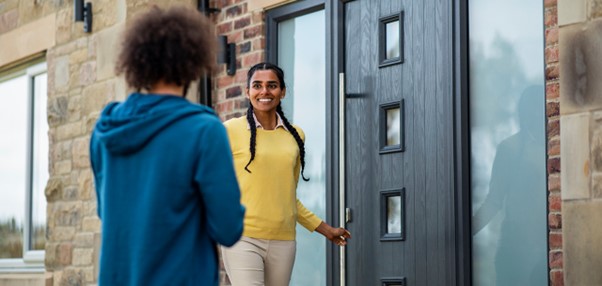 Smiling woman with braids in yellow top opens a dark grey composite front door, greeting a person in a blue hoodie.