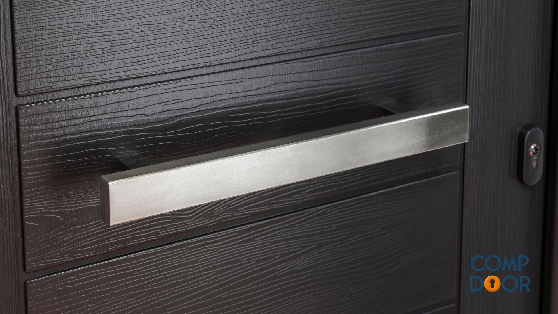 Black composite composite door with horizontal wood grain texture, long silver brushed handle, and keyhole. Blue COMP door logo.