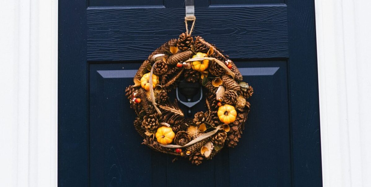 Autumn wreath with pinecones, orange mini pumpkins, red berries, on a dark blue composite door with white trim.