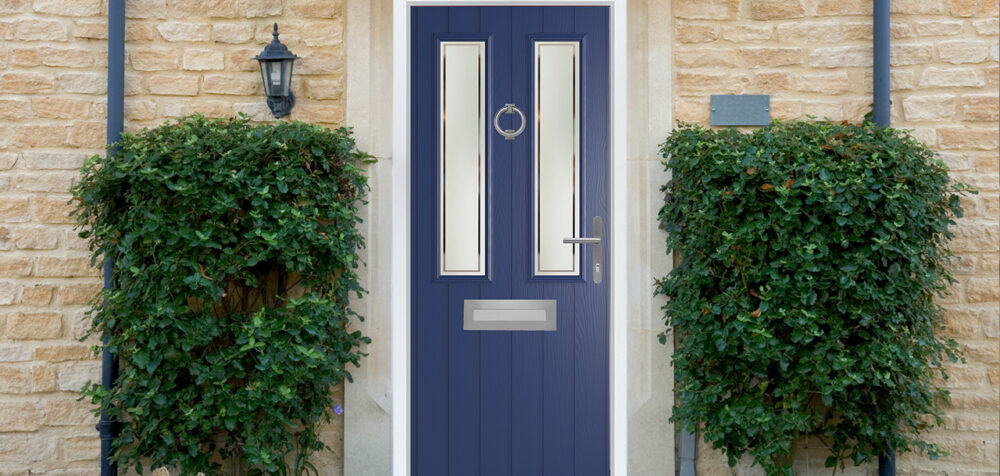 White-framed dark blue composite door with 2 glass panels, silver knocker, handle, letterbox, on a light brick wall with green bushes.